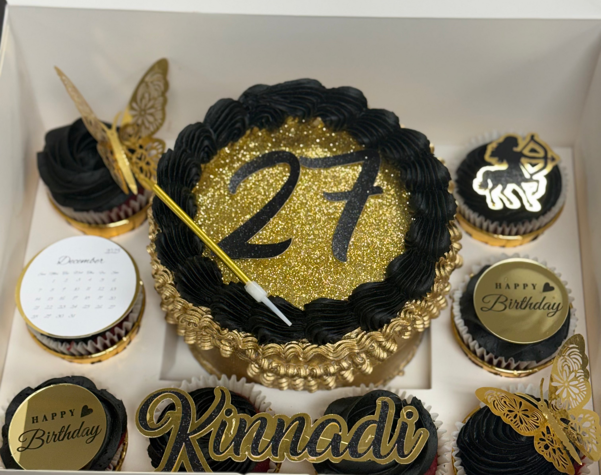 Birthday cake with number 27 and name 'Kinnadi' surrounded by cupcakes in a box.